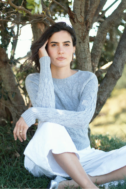 Woman wearing a light grey sweater and white pants sitting outdoors. #colour_light-grey